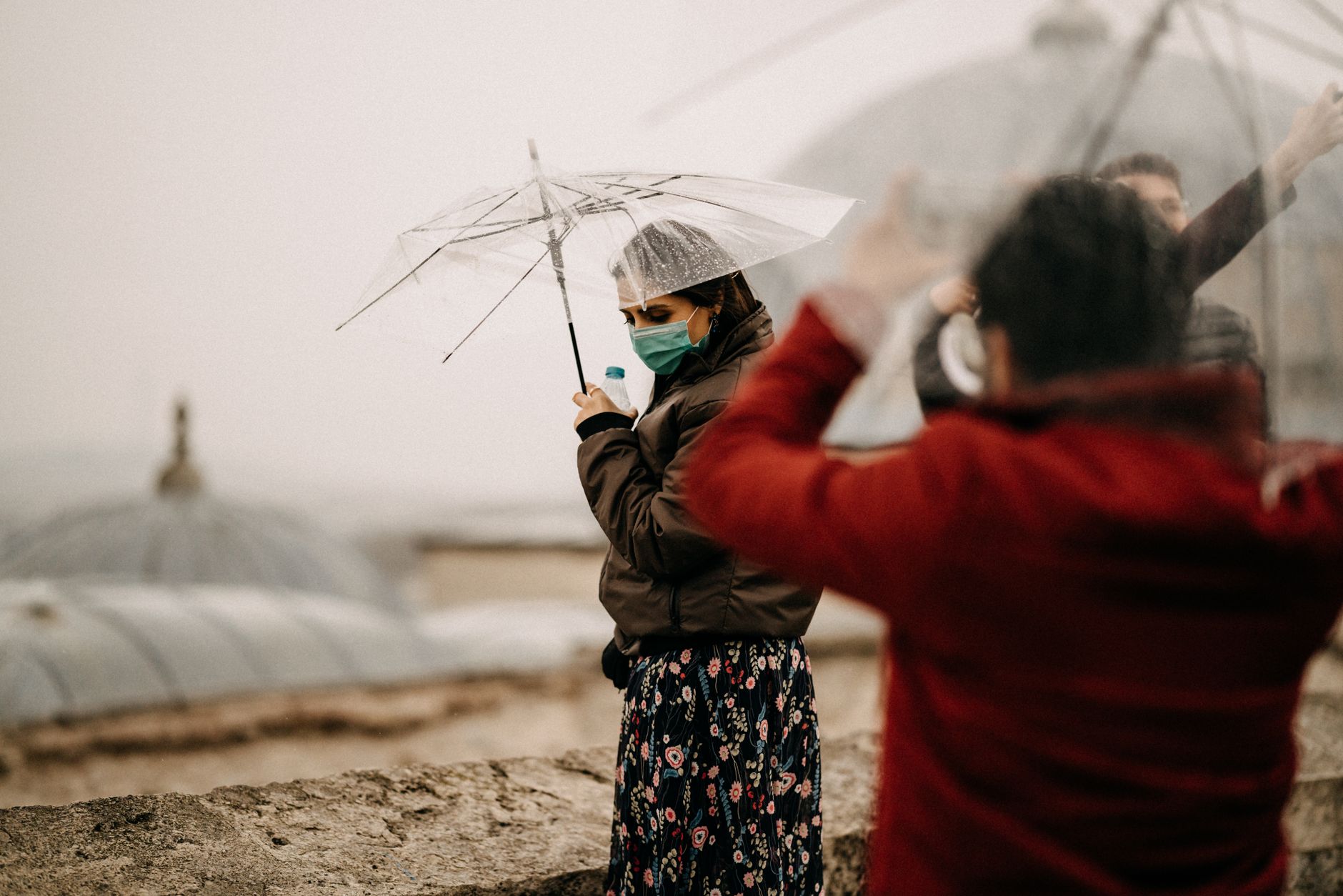 Woman wearing face mask and holding transparent umbrella on a rainy city landscape.